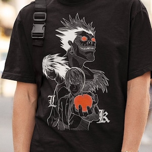May include: Black t-shirt featuring a graphic design with three characters and the letters "L" and "K". The central figure holds a red apple. The design is in white and gray, with red accents. The t-shirt is short-sleeved.