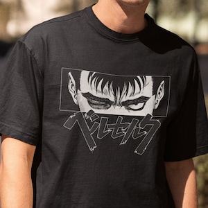 May include: Black t-shirt featuring a manga-style graphic of a character's eyes and the Japanese text "ベルセルク" in white. The short-sleeved shirt is made of a soft material and is suitable for casual wear.