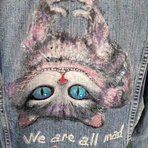 Könnte beinhalten: Eine blaue Jeansjacke mit einer handgemalten Illustration einer auf dem Kopf stehenden Katze mit leuchtend blauen Augen und rosa Akzenten. Die Katze formt mit ihren Pfoten ein Herz über ihrem Kopf. Unter der Katze steht der Text, We are all mad in love here.
