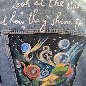 Puede incluir: Chaqueta vaquera azul claro con texto bordado en blanco: "Look at the stars, look how they shine for". La parte trasera presenta una escena nocturna pintada con estrellas, planetas y formas arremolinadas.