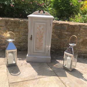 May include: A grey cabinet with a floral design on the door is flanked by two silver lanterns with candles. The cabinet has a decorative top and a small door handle. The lanterns have a handle at the top and are made of metal and glass.