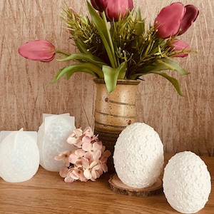 Könnte beinhalten: Vier weiße Keramikeier mit strukturierter Oberfläche, auf einem Holztisch angeordnet. Eine Vase mit rosa Blumen steht im Hintergrund.