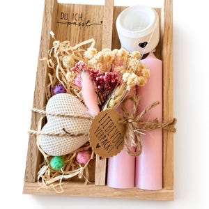 May include: A wooden gift box containing two light pink pillar candles, a white heart-shaped textured object, a white candle holder with a black heart, and a dried flower bouquet. A brown paper tag reads LIEBEVOLL VERPACKT and text reads DU ICH passt.