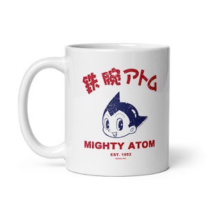 May include: White ceramic mug with a blue and black illustration of Astro Boy, the Japanese manga character. The mug has red text that says "Mighty Atom Est. 1952 Tezuka Inc."