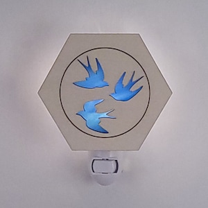 May include: A white hexagonal nightlight with a blue silhouette of three birds flying in a circle. The nightlight has a standard plug-in base.