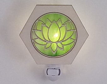 以下が含まれることがあります： 緑の蓮の花のデザインが施された白い六角形のナイトライト。ライトは点灯しており、花を照らしています。