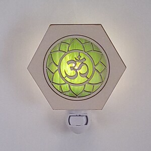 Könnte beinhalten: Ein weißes sechseckiges Nachtlicht mit einem grünen und weißen Om-Symbol in einem Lotusblüten-Design. Das Nachtlicht hat einen weißen Steckerfuß.