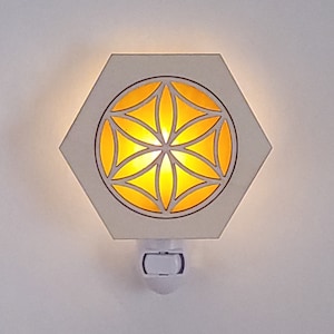 May include: White hexagonal night light with a cut-out design of a flower of life in the center. The light is on and illuminates the design in a warm yellow color.