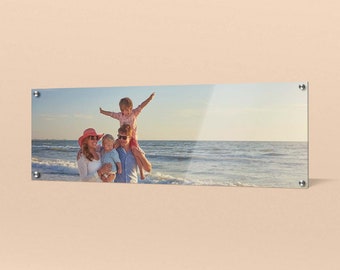 Op de afbeelding: Een gezin van vier op een strand, de ouders houden hun twee kinderen vast. De vader draagt ​​het oudste kind op zijn schouders, terwijl de moeder het jongste kind in haar armen houdt. Het gezin glimlacht en kijkt naar de camera. Het strand is zanderig en het water is blauw.