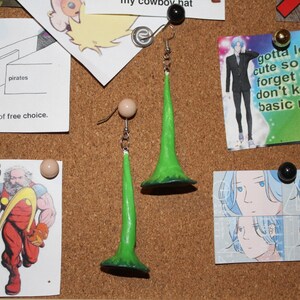 May include: A pair of green and silver earrings with a long, pointed, green, horn-like design. The earrings are hanging on a cork board.