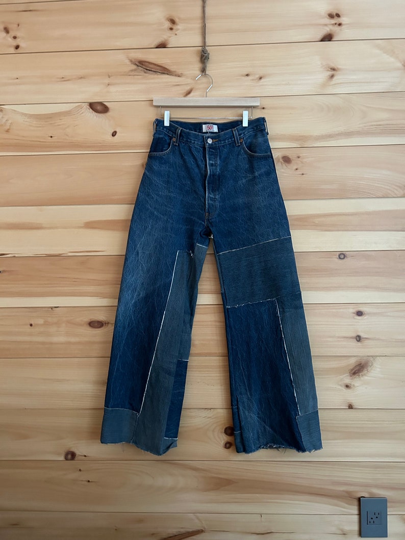 以下が含まれることがあります： パッチワークのディテールとワイドレッグが特徴のブルーデニムジーンズ。ジーンズは木製のハンガーに吊るされ、木製のパネルの壁を背景にしています。ジーンズはボタン留めで、501のロゴが付いています。