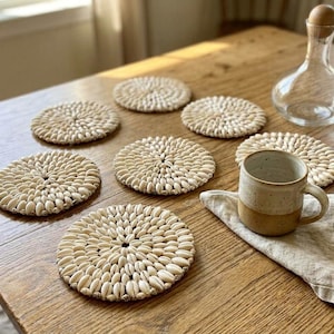 May include: A set of seven circular coasters crafted from small, off-white cowrie shells arranged in a radial pattern. Each coaster features a consistent, textured design with the shells tightly packed together to form a flat, round surface.