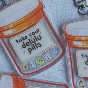 Könnte beinhalten: Transparente Acryl-Schlüsselanhänger in Form von orangefarbenen Medikamentendosen mit weißen Deckeln und silbernen Karabinerverschlüssen. Jedes Etikett trägt den Text "take your delulu pills" und kleine, bunte Cartoon-Figuren-Symbole.