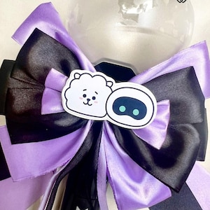 Op de afbeelding: Een helder, bolvormig object is versierd met een zwart en paars lint. De strik heeft een zwart midden en paarse details, met een sticker van een wit cartoon schaap en een blauwogig karakter. Het object heeft een zwarte handgreep met een logo.