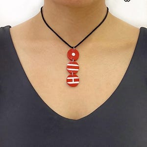 以下が含まれることがあります： 3つの円形チャームが付いた赤いペンダントネックレス。一番上のチャームには白い点があり、他の2つには白い横縞があります。ネックレスは黒いコードから吊り下げられています。背景は濃い灰色のトップスです。