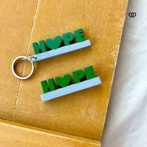 May include: Two green wooden keychains with the word "HOPE" and a heart in the middle. The keychains are on a brown surface.