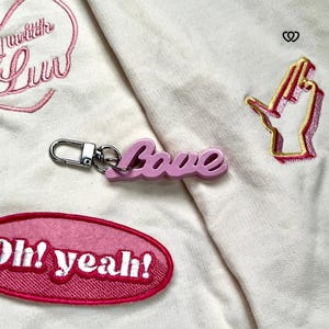 May include: Pink keyring with a silver ring and the word "love" in pink 3D letters. The keyring is on a white fabric background with pink embroidered patches that say "with the luv" and "Oh! yeah!"