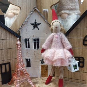 Peut inclure: Une poupée en tricot rose portant un bonnet de Père Noël rouge et tenant une lanterne blanche. La poupée est debout devant une maison miniature en bois avec une étoile sur le toit. Il y a une tour Eiffel rose au premier plan.