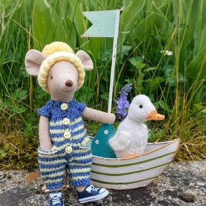 May include: A knitted mouse toy in a blue and yellow striped outfit, a yellow hat, and blue trainers. Next to the mouse is a white knitted duck in a small boat with a green and white striped design and a small flag.