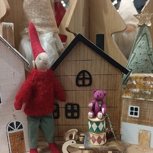Peut inclure: Un lutin en bois rouge et vert avec un chapeau rose se tient à côté d'une maison en bois avec un ours en peluche violet assis sur une pile de boîtes colorées dans un traîneau en bois.