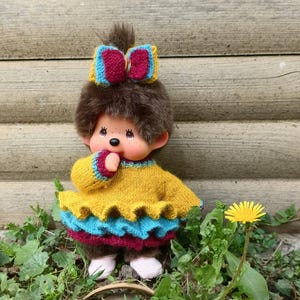 May include: A plush doll with brown fur, wearing a yellow sweater with blue and maroon trim. The doll has a matching bow and is standing in grass. The background is a wooden fence.