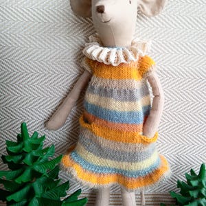 Peut inclure: Une souris en peluche portant une robe tricotée rayée avec un col en crochet blanc. La souris est debout sur une surface texturée avec deux arbres jouets verts.