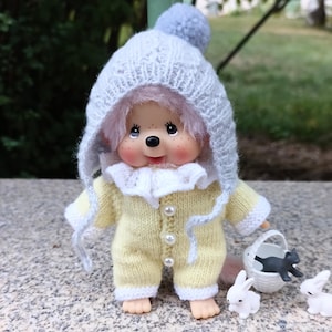 May include: A doll wearing a light yellow knitted jumpsuit with white trim and pearl buttons. The doll has a white knitted hood with a blue pom-pom. A small white basket holds a black cat figurine, with two white bunny figurines nearby.
