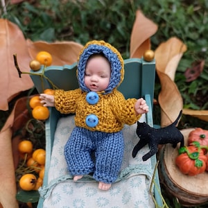 Peut inclure: Une poupée miniature au teint pâle portant un pull en tricot à capuche jaune moutarde avec deux boutons bleus et un pantalon en tricot bleu assorti, allongée dans un petit lit en bois bleu clair avec un matelas en tissu blanc et bleu.