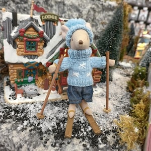 May include: A small, plush mouse wearing a blue knitted sweater with white snowflakes and blue jeans is skiing down a snowy mountain. The mouse is holding ski poles and is in front of a small wooden lodge with a sign that reads "LODGE".
