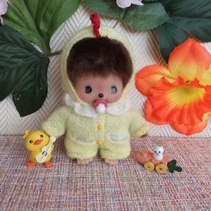 May include: A small doll wearing a yellow knitted outfit with a hood and a pacifier. A yellow toy chick with a guitar and a wooden toy with a bunny are also present. The doll has brown fur and a red comb.