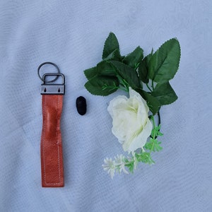 May include: A brown leather keyring with a silver metal ring and two silver metal loops. The keyring is on a white background with a white rose and green leaves.