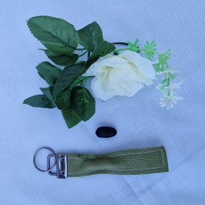 May include: A green leather keychain with a silver key ring and a white rose with green leaves in the background.