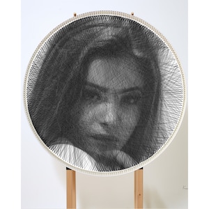 Puede incluir: Un retrato en blanco y negro de la cara de una persona creado con arte de hilo. El arte de hilo está montado en un marco redondo de madera con un fondo blanco.