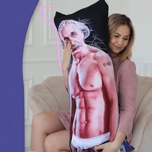 May include: A body pillow featuring a muscular illustration of Albert Einstein. The pillow has a black background with scientific equations and a portrait of Einstein. The pillow is being held by a person wearing a pink shirt.