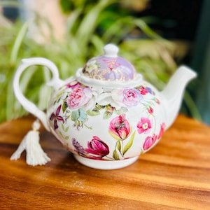 May include: A white porcelain teapot with a floral design featuring pink roses, purple tulips, and green leaves. The teapot has a tassel on the spout and a lid with a purple and gold floral design.