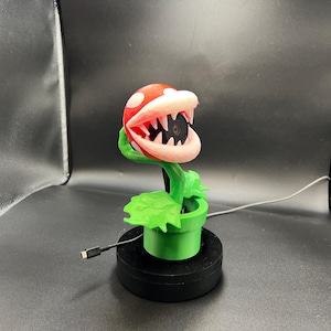 May include: A 3D-printed Piranha Plant from the Super Mario video game series. The plant has a red head with white spots and a white-and-pink mouth filled with teeth. It sits in a green pot on a black base, with a USB cable attached.
