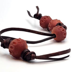 May include: A close-up of a bracelet featuring reddish-brown, textured beads and dark brown leather cords. The beads have a unique, pitted surface. The leather cords are knotted, creating a decorative and functional design.