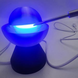 May include: A spherical lamp with a dark base and a glowing blue upper section, featuring a white light emitting from the center gap. A silver tool is inserted into the opening between the two halves, and a white USB power cable is attached.