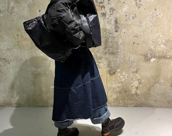 May include: A person wearing a black puffer jacket and a long denim skirt, carrying a large black tote bag. The person is wearing black boots with orange accents.