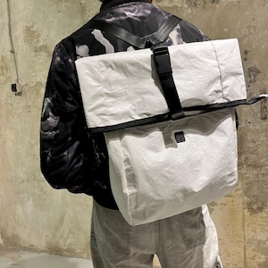Puede incluir: Una mochila blanca de tela arrugada con una correa y una hebilla negras. La mochila la lleva puesta una persona que lleva una chaqueta con estampado blanco y negro.