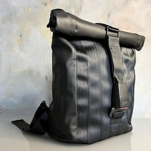 May include: Black roll-top backpack made from repurposed seatbelts. The bag features a vertical striped pattern, a buckle closure, and a shoulder strap. The top is rolled down and secured with a strap.