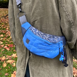 Può includere: Una borsa a tracolla trapuntata blu con un pannello superiore a motivo floreale blu scuro, una tracolla grigia regolabile e una piccola toppa con logo a forma di volpe nera. La borsa presenta uno scomparto principale con cerniera.