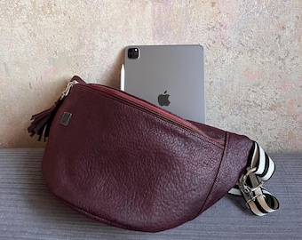 May include: A crescent-shaped burgundy crossbody bag with a textured surface, a zipper closure, and a tassel pull. The bag features a black and white striped adjustable strap with silver-toned metal hardware and a small square logo patch.