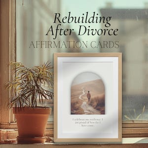 May include: A framed print with the text "Rebuilding After Divorce AFFIRMATION CARDS." The print features an image of a person walking on a path. The text "I celebrate my resilience. I am proud of how far I have come." is below the image. A potted plant sits in front of a window.