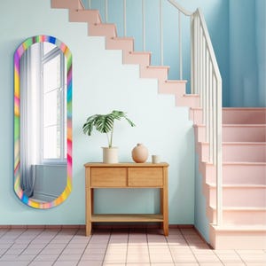 Op de afbeelding: Een hoge, ovaalvormige wandspiegel met een levendig, meerkleurig regenboogverloopframe. De spiegel reflecteert een raam met witte gordijnen, terwijl het frame een kleurenspectrum vertoont met rood, oranje, geel, groen, blauw en paars.