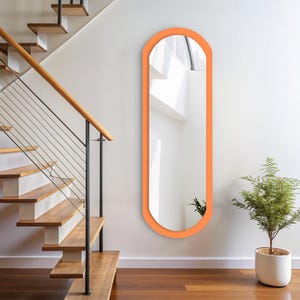 Op de afbeelding: Een hoge passpiegel met een glad, aan de bovenkant afgerond frame in een levendige oranje kleur. De spiegel heeft een verticale, langwerpige ovale vorm en is plat tegen een effen witte muur gemonteerd, wat licht en ruimte reflecteert.