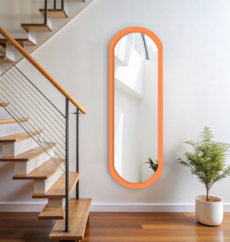 Op de afbeelding: Een hoge passpiegel met een glad, aan de bovenkant afgerond frame in een levendige oranje kleur. De spiegel heeft een verticale, langwerpige ovale vorm en is plat tegen een effen witte muur gemonteerd, wat licht en ruimte reflecteert.