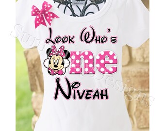 May include: White t-shirt with a pink and white polka dot Minnie Mouse design. The shirt says "Look Who's One" with the word "one" in pink polka dots. The name "Niveah" is written in black below the design.
