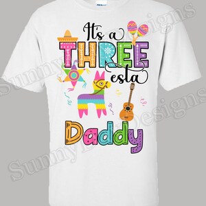May include: White t-shirt with a colorful graphic design that says "It's a Three esta Daddy". The design includes a pinata, a guitar, and other festive decorations.