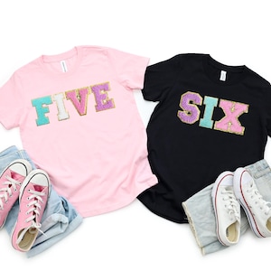 May include: Two t-shirts with the words "FIVE" and "SIX" in colorful, glittery letters. The shirts are pink and black and are laid out on a white background with a pair of white sneakers and blue jeans.